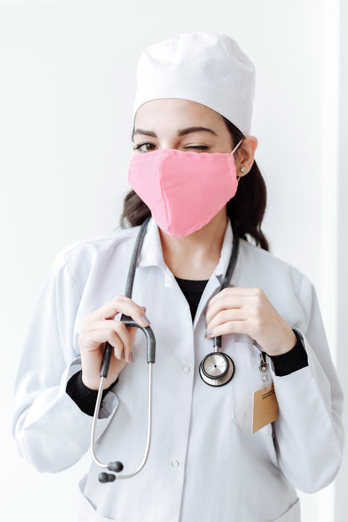 Female nurse winking in white coat with stethoscope and pink mask, indoors.